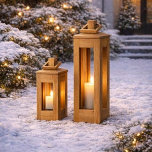 May include: Two wooden lanterns with lit candles sit in the snow. The lanterns are rectangular with open sides, and a handle on top. The scene is outdoors, with snow-covered bushes and a house in the background. The warm glow of the candles contrasts with the cold winter scene.