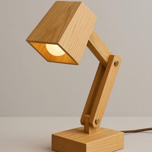 May include: A wooden desk lamp with a square base and a rectangular shade. The lamp is made of light-coloured wood and features adjustable arms. The light bulb is visible inside the shade. The lamp is connected to a brown cord.