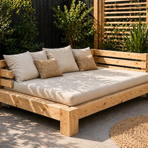 Könnte beinhalten: Ein helles Outdoor-Tagesbett aus Holz mit einem beigen Kissen und mehreren beigen und strukturierten Zierkissen. Das Tagesbett hat eine Lattenlehne und Armlehne. Die Szene spielt sich im Freien mit Grün und einer gewebten Matte ab.