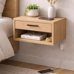 May include: A floating wooden nightstand with a drawer and open shelf. The nightstand has a black handle, a book, glasses, and a small woven basket on the shelf. A white vase with dried flowers, a small plant, and a smartphone are on top. A phone is charging on the floor.