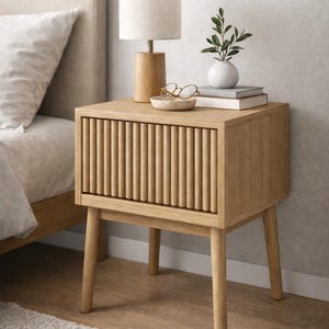 Puede incluir: Una mesita de noche de roble claro con un frente de cajón con lamas verticales. La mesa tiene una lámpara pequeña, un jarrón con vegetación, libros y un cuenco pequeño con gafas encima. La mesa tiene cuatro patas cónicas.