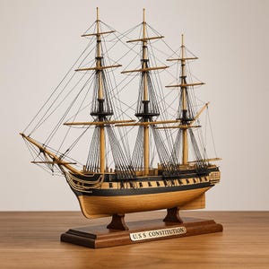 May include: A detailed wooden model of the U.S.S. Constitution, a three-masted sailing ship. The model features tan masts, black rigging, and a dark wooden hull. The ship rests on a wooden base with a nameplate that reads "U.S.S. Constitution."