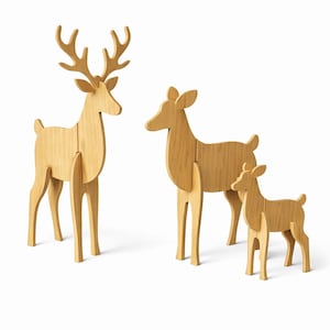 Può includere: Tre sculture di cervi in legno di diverse dimensioni. Il più grande ha le corna, gli altri no. Sono realizzati in legno chiaro e si ergono su uno sfondo bianco. Il cervo più alto misura circa 30 cm.