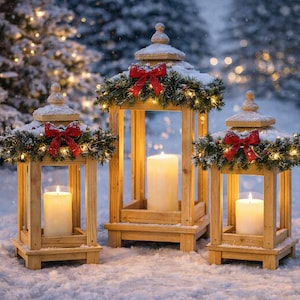 Puede incluir: Tres faroles de madera con velas encendidas, decorados con ramas de hoja perenne, lazos rojos y luces de hadas, están en la nieve. Los faroles son de diferentes tamaños y tienen un diseño clásico y festivo, ideal para la decoración navideña.