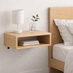May include: A light wood floating nightstand with a white lamp, a glass of water with a sprig of greenery, and two books. The nightstand is mounted on a white wall next to a bed with white bedding.