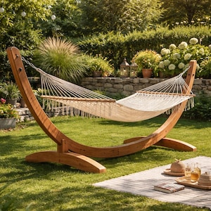 Könnte beinhalten: Eine Holz-Hängematte mit cremefarbenem Webbett und Fransen hängt zwischen zwei gebogenen Holzstützen. Die Hängematte steht auf einem grünen Rasen in einem Garten. Eine Picknickdecke, Getränke und ein Hut liegen auf dem Gras.