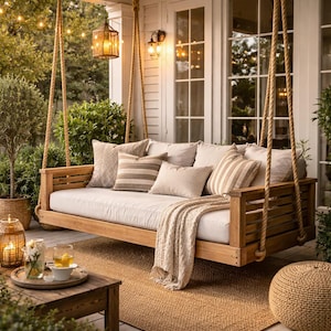 May include: A wooden porch swing with beige cushions and pillows hangs from thick ropes. A beige throw blanket and a woven rug add to the cozy outdoor seating area. Lanterns and string lights create a warm ambiance.