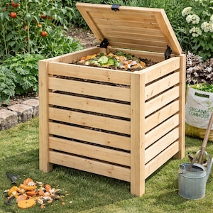 Op de afbeelding: Een houten compostbak met een open deksel, gevuld met organisch materiaal. De bak is gemaakt van lichtgekleurd hout en heeft een lattenontwerp. Hij staat op een grasveld, met een tuin en planten op de achtergrond. Een zak met organische compost is zichtbaar.