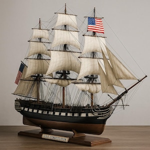 Könnte beinhalten: Ein detailliertes Modell der U.S.S. Constitution, einem Dreimast-Segelschiff. Das Schiff hat weiße Segel, einen schwarzen und braunen Rumpf und amerikanische Flaggen. Das Modell ist auf einem Holzsockel mit einem Namensschild "U.S.S. Constitution" montiert.