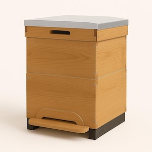 Puede incluir: Una colmena de madera con una tapa gris claro y una base oscura. La colmena está construida con madera de color marrón claro y presenta un asa en la parte delantera. El diseño es simple y funcional, adecuado para la apicultura.
