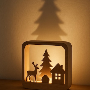 May include: Wooden Christmas-themed night light with a deer, tree, and house silhouette. The warm light casts a tree shadow on the wall. The lamp is made of light-colored wood and sits on a wooden surface.