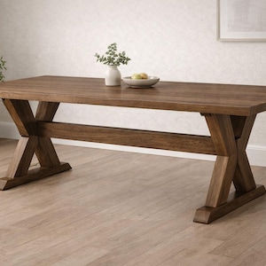 May include: A large, rectangular wooden dining table with a dark brown finish. The table features an X-shaped base for support. A small vase with greenery and a bowl of fruit are on the tabletop. The table is approximately 72 inches long.