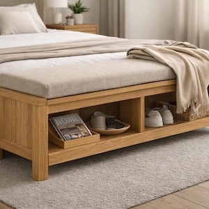 Może przedstawiać: Drewniana ławka do przechowywania z jasnobeżową poduszką i otwartymi półkami. Ławka znajduje się u podnóża łóżka z pościelą w neutralnych odcieniach. W przegrodach do przechowywania widoczny jest magazyn, buty i kosz. Ławka ma około 150 cm długości.