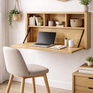 May include: A light oak wall-mounted desk with a drop-down work surface, a laptop, and a chair. The desk features shelves with books, plants, and a woven basket. A round mirror and a hanging plant are also visible.