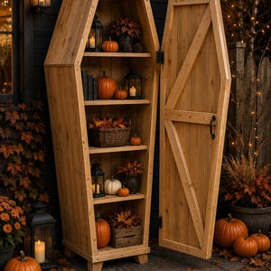 May include: A tall, light-brown wooden cabinet shaped like a coffin with an open door. The interior features four shelves holding various items including orange pumpkins, black metal lanterns with candles, a wicker basket, and autumn leaves.