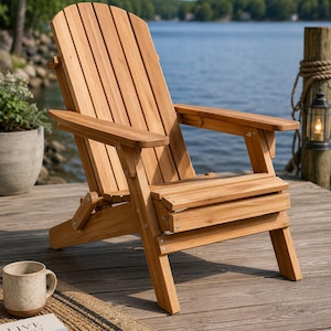 Könnte beinhalten: Ein hölzerner Adirondack-Stuhl mit natürlichem Finish steht auf einem Holzsteg mit Blick auf einen See. Der Stuhl hat eine Lattenlehne und -sitzfläche mit breiten Armlehnen. Der Stuhl ist zusammengeklappt. Eine Tasse und ein Buch sind im Vordergrund.