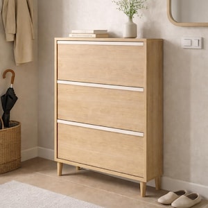 Può includere: Un mobile porta scarpe in legno chiaro con tre cassetti e dettagli bianchi. Il mobile è in un corridoio con un tappeto beige, un cappotto e un ombrello. Sono visibili anche un vaso con fiori e uno specchio.