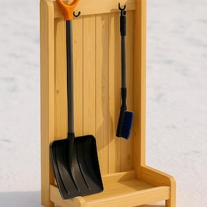May include: A wooden snow tool organizer with a black snow shovel and a black brush with blue bristles. The shovel has an orange handle. The organizer is made of light-colored wood and has a small shelf at the bottom.
