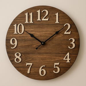 Könnte beinhalten: Eine runde Wanduhr aus Holz mit dunkelbraunem Finish. Das Zifferblatt hat große, hellfarbene Ziffern und schwarze Zeiger. Das Design der Uhr ist schlicht und rustikal, passend für die Inneneinrichtung.
