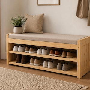 May include: A wooden shoe rack with two shelves, holding various pairs of shoes. The rack has a padded seat cushion and is placed against a neutral-colored wall. A small potted plant and a decorative pillow are on top.