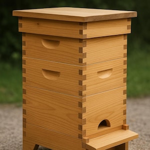 May include: A wooden beehive with a natural finish. The hive has a rectangular shape with multiple stacked boxes, each with a recessed handle. A small landing platform extends from the front. The hive is constructed with dovetail joints.