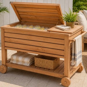 May include: A wooden outdoor cooler with a hinged lid, filled with ice and drinks. It has a lower shelf with a basket and folded towels. The cooler is on wheels and has a side table with a towel rack. A fern in a basket sits on top.