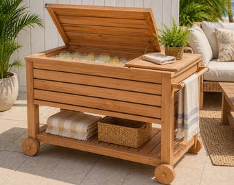 Pode incluir: Um refrigerador de exterior de madeira com tampa articulada, cheio de gelo e bebidas. Possui uma prateleira inferior com uma cesta e toalhas dobradas. O refrigerador está sobre rodas e tem uma mesa lateral com um toalheiro.