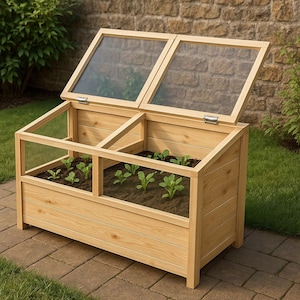 May include: A wooden cold frame with a hinged, clear-paneled lid. The rectangular frame is filled with soil and small green plants. The cold frame is made of light-coloured wood and sits on a brick patio, with greenery in the background.