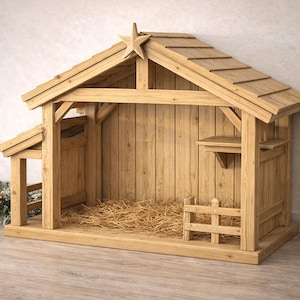 May include: A wooden nativity stable with a star on the roof. The structure is made of light-colored wood and features a sloped roof, a small fence, and a hay-filled interior. A Christmas tree is in the background.