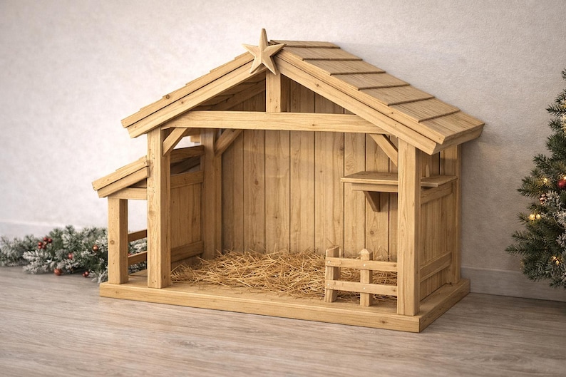 May include: A wooden nativity stable with a star on the roof. The structure is made of light-colored wood and features a sloped roof, a small fence, and a hay-filled interior. A Christmas tree is in the background.