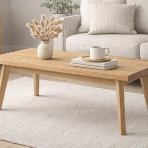 May include: A light-colored wooden coffee table with angled legs. A white mug sits on a wooden tray atop a stack of books. A vase with dried flowers is on the table. A light-colored rug is beneath the table.