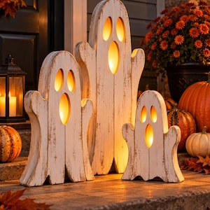 May include: Three white wooden ghost decorations with a distressed finish, each featuring cut-out eyes and a mouth. The hollow centers are illuminated from within by a warm, glowing light, creating a bright, soft radiance through the openings.