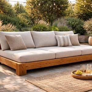 以下が含まれることがあります： ベージュのクッションと枕が付いた木製のアウトドアデイベッド。 デイベッドは、織りのラグのある石のパティオにあります。 花瓶に入った花と飲み物と果物のトレイが付いた小さなサイドテーブルが近くにあります。 背景には緑豊かな緑があります。