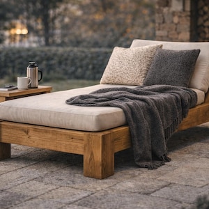 May include: A wooden outdoor chaise longue with a cream cushion, two pillows, and a grey blanket. A small side table holds a mug and a flask. A lit candle in a lantern adds ambiance.