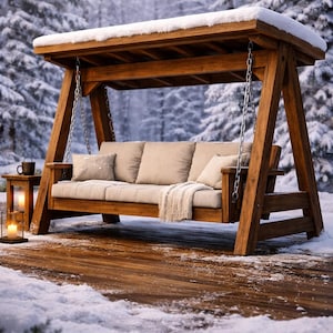 Könnte beinhalten: Eine Holzschaukel mit Baldachin, Kissen und einer Decke, die sich im Freien in einer verschneiten Umgebung befindet. Die Schaukel ist aus hellbraunem Holz gefertigt und hängt an Ketten. Ein kleiner Beistelltisch mit einer Tasse und Kerzen steht in der Nähe.
