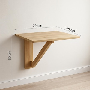 Puede incluir: Mesa de madera clara montada en la pared. La encimera rectangular mide 70 cm de ancho y 40 cm de profundidad, sostenida por un soporte diagonal y un soporte de pared vertical. La mesa tiene 50 cm de altura.