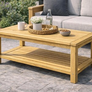 May include: A rectangular wooden coffee table with a lower shelf. The table is light brown and has a slatted top and shelf. A white mug, a woven tray with flowers, a bowl of nuts, and a water bottle are on the table. The table is outdoors.