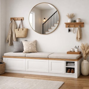 May include: A contemporary hallway scene with a white storage bench, light beige cushion, and wooden detailing. A round gold mirror is above the bench, with a wooden coat rack and shelf on either side. Cushions and a woven bag complete the decor.