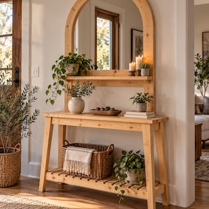 May include: A light-colored wooden console table with a matching arched mirror attached to the back. The unit features a top shelf, a main surface, and a slatted bottom shelf. It is decorated with potted plants, candles, a wooden bowl, and a woven basket.