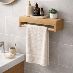 May include: A wooden bathroom shelf with a towel bar, holding a cream-colored towel. The shelf displays a soap dispenser, bottles, a small plant, and a tray. The background is a tiled wall with a round mirror.