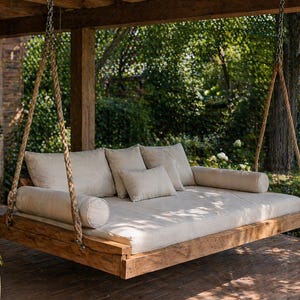 May include: A wooden swing bed with a light-colored mattress and pillows hangs from a porch. The swing is suspended by thick ropes and chains. The background features lush greenery and a wooden structure.