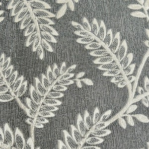 K&ouml;nnte beinhalten: Nahaufnahme eines grauen Stoffes mit einem wei&szlig;en, gestickten Blumenmuster. Das Muster zeigt zarte Bl&auml;tter und Zweige.