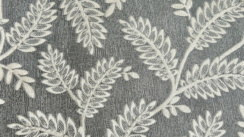K&ouml;nnte beinhalten: Nahaufnahme eines grauen Stoffes mit einem wei&szlig;en, gestickten Blumenmuster. Das Muster zeigt zarte Bl&auml;tter und Zweige.