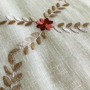 Puede incluir: Un mantel de tela blanco con un delicado dise&ntilde;o bordado de hojas marrones y flores rojas.