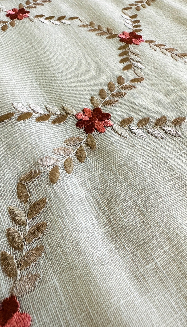 Puede incluir: Un mantel de tela blanco con un delicado dise&ntilde;o bordado de hojas marrones y flores rojas.