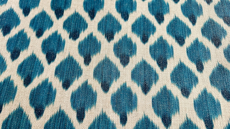 May include: A close-up of a fabric with a blue and white ikat pattern. The fabric has a textured surface and a repeating pattern of blue teardrop shapes on a white background.