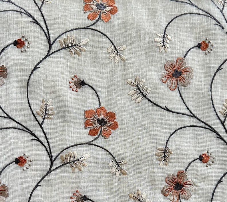 K&ouml;nnte beinhalten: Ein beigefarbener Stoff mit gestickten Blumendesigns in Schwarz und Orange. Die Blumen sind stilisiert und haben ein einfaches Design.