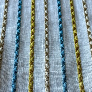 May include: Close-up of a white fabric with embroidered stripes in blue, yellow, and beige.