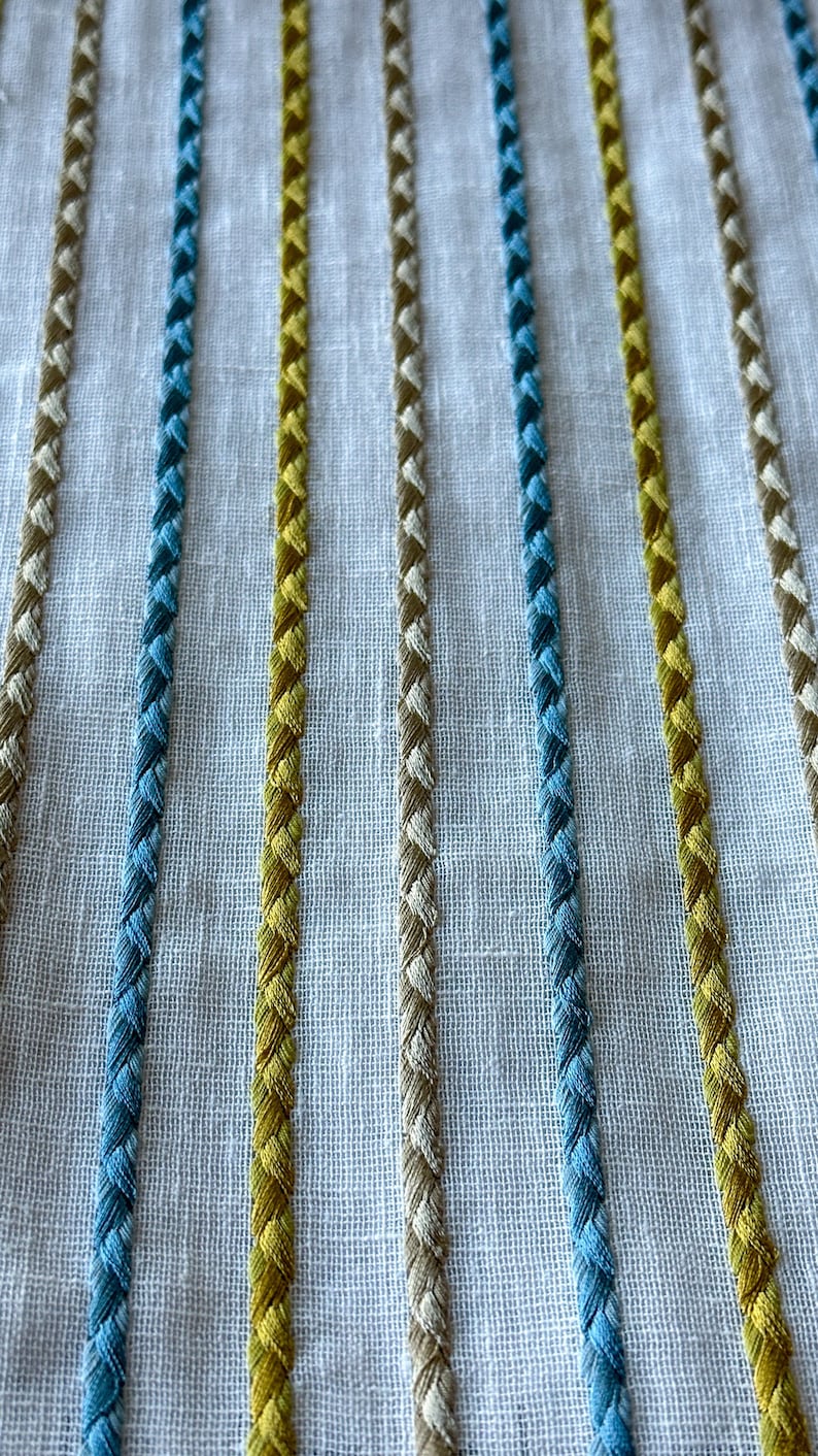 May include: Close-up of a white fabric with embroidered stripes in blue, yellow, and beige.