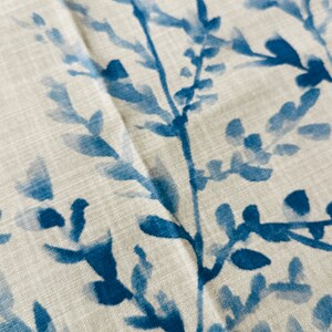 May include: Close-up of a fabric with a watercolor-style botanical print. The design features blue branches and leaves against a neutral, off-white background. The fabric has a textured appearance, suggesting a linen or cotton blend.